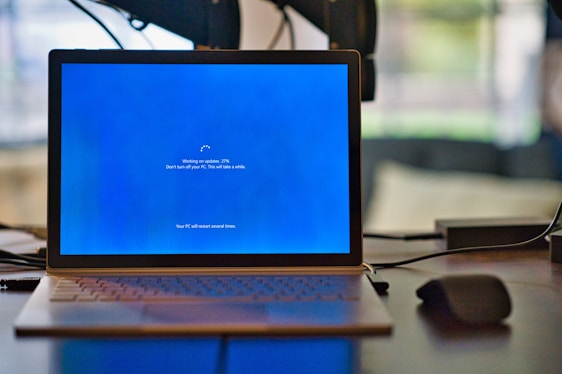 A laptop on a desk is displaying a blue screen with an update notification. The screen shows a progress indicator for Windows updates at 27%, along with text advising not to turn off the PC. A mouse is placed next to the laptop on the desk.
