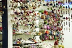 A store display filled with an assortment of colorful handcrafted items including plush toys, hanging decorations, and patterned pouches. Shelves are packed with creatively designed products, exhibiting vibrant textiles and intricate patterns. The scene is lively with a clutter of colors and textures, giving a sense of artisanal craftsmanship.