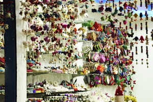 A store display filled with an assortment of colorful handcrafted items including plush toys, hanging decorations, and patterned pouches. Shelves are packed with creatively designed products, exhibiting vibrant textiles and intricate patterns. The scene is lively with a clutter of colors and textures, giving a sense of artisanal craftsmanship.