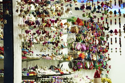 A store display filled with an assortment of colorful handcrafted items including plush toys, hanging decorations, and patterned pouches. Shelves are packed with creatively designed products, exhibiting vibrant textiles and intricate patterns. The scene is lively with a clutter of colors and textures, giving a sense of artisanal craftsmanship.
