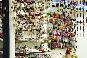 A store display filled with an assortment of colorful handcrafted items including plush toys, hanging decorations, and patterned pouches. Shelves are packed with creatively designed products, exhibiting vibrant textiles and intricate patterns. The scene is lively with a clutter of colors and textures, giving a sense of artisanal craftsmanship.