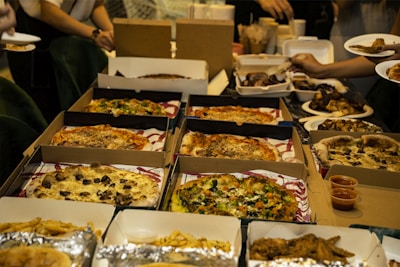 An assortment of pizzas and fried foods is displayed on a table. Several pizza boxes are open, revealing various toppings such as mushrooms, vegetables, and cheese. There are also containers with fried chicken and plates with food being held by people reaching for servings.