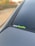 A close-up view of a car's rear window featuring a sticker with the text 'CarMeets'. The sticker has a combination of white and green lettering. The background is slightly blurred, with hints of greenery and a touch of autumn colors.