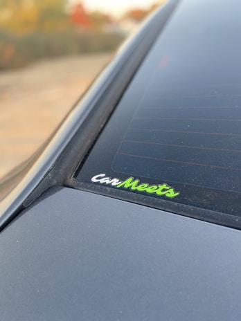 A close-up view of a car's rear window featuring a sticker with the text 'CarMeets'. The sticker has a combination of white and green lettering. The background is slightly blurred, with hints of greenery and a touch of autumn colors.