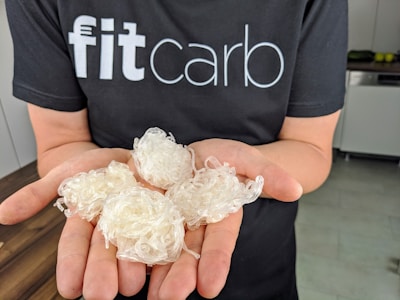 A person wearing a black t-shirt with the text 'fitcarb' holding several white, translucent coiled objects in their palms. The background features a kitchen setting with a wooden countertop and some kitchen appliances.
