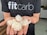 A person wearing a black t-shirt with the text 'fitcarb' holding several white, translucent coiled objects in their palms. The background features a kitchen setting with a wooden countertop and some kitchen appliances.