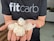 A person wearing a black t-shirt with the text 'fitcarb' holding several white, translucent coiled objects in their palms. The background features a kitchen setting with a wooden countertop and some kitchen appliances.