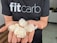 A person wearing a black t-shirt with the text 'fitcarb' holding several white, translucent coiled objects in their palms. The background features a kitchen setting with a wooden countertop and some kitchen appliances.