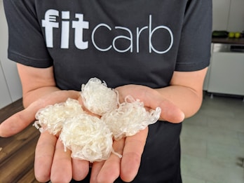 A person wearing a black t-shirt with the text 'fitcarb' holding several white, translucent coiled objects in their palms. The background features a kitchen setting with a wooden countertop and some kitchen appliances.
