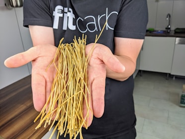 A person is holding a bunch of thin, dried noodles in their open palms. They are wearing a dark t-shirt with partially visible text in a kitchen setting.