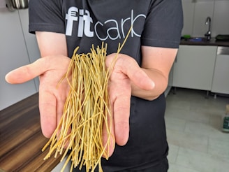 A person is holding a bunch of thin, dried noodles in their open palms. They are wearing a dark t-shirt with partially visible text in a kitchen setting.