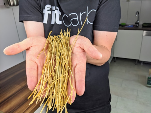A person is holding a bunch of thin, dried noodles in their open palms. They are wearing a dark t-shirt with partially visible text in a kitchen setting.