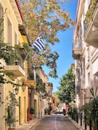 A picturesque street lined with colorful buildings and lush greenery, featuring a Greek flag waving gently in the breeze. The street is narrow and inviting, showcasing traditional architecture with balconies and quaint street lamps. The vibrant vegetation and clear blue sky add a refreshing and cheerful element to the scene.