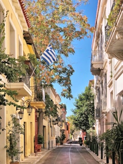 A picturesque street lined with colorful buildings and lush greenery, featuring a Greek flag waving gently in the breeze. The street is narrow and inviting, showcasing traditional architecture with balconies and quaint street lamps. The vibrant vegetation and clear blue sky add a refreshing and cheerful element to the scene.