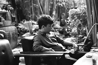 A young person sits in a contemplative pose in a lush, plant-filled garden. The monochrome setting includes a large decorative pot on the left and various plants around. The person is relaxed, leaning back in a chair with one hand resting in their lap and the other on the armrest. The scene is peaceful and intimate.
