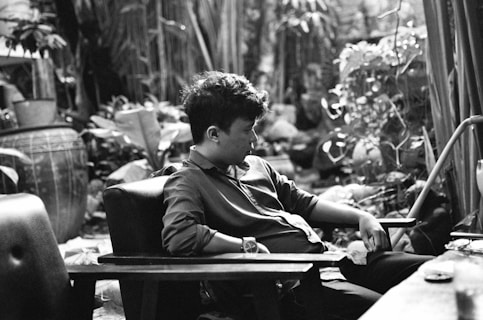 A young person sits in a contemplative pose in a lush, plant-filled garden. The monochrome setting includes a large decorative pot on the left and various plants around. The person is relaxed, leaning back in a chair with one hand resting in their lap and the other on the armrest. The scene is peaceful and intimate.