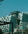 A modern, uniquely designed building with multiple reflective glass panels that create a checkered pattern. The upper parts display the names 'Norton Rose Fulbright' and another in Chinese characters. The structure exhibits a futuristic architectural style with sharp angles and a mix of straight and curved lines. In the foreground, additional buildings are visible with balconies and greenery, suggesting an urban setting.