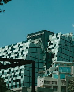 A modern, uniquely designed building with multiple reflective glass panels that create a checkered pattern. The upper parts display the names 'Norton Rose Fulbright' and another in Chinese characters. The structure exhibits a futuristic architectural style with sharp angles and a mix of straight and curved lines. In the foreground, additional buildings are visible with balconies and greenery, suggesting an urban setting.