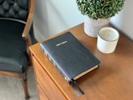 A black leather-bound Bible rests on a wooden table alongside a small potted plant with green foliage and a white candle in a ceramic holder. In the background, the edge of a black cushioned chair is visible.