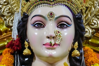 A detailed and intricately crafted sculpture of a deity adorned with elaborate jewelry, including golden earrings and a nose ring embellished with pearls and gemstones. The figure has strikingly painted eyes and a serene expression. The headdress is ornate, featuring gold and colored stones, and is surrounded by marigold flowers.