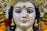 A detailed and intricately crafted sculpture of a deity adorned with elaborate jewelry, including golden earrings and a nose ring embellished with pearls and gemstones. The figure has strikingly painted eyes and a serene expression. The headdress is ornate, featuring gold and colored stones, and is surrounded by marigold flowers.