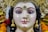 A detailed and intricately crafted sculpture of a deity adorned with elaborate jewelry, including golden earrings and a nose ring embellished with pearls and gemstones. The figure has strikingly painted eyes and a serene expression. The headdress is ornate, featuring gold and colored stones, and is surrounded by marigold flowers.