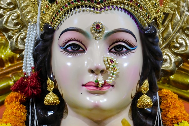 A detailed and intricately crafted sculpture of a deity adorned with elaborate jewelry, including golden earrings and a nose ring embellished with pearls and gemstones. The figure has strikingly painted eyes and a serene expression. The headdress is ornate, featuring gold and colored stones, and is surrounded by marigold flowers.