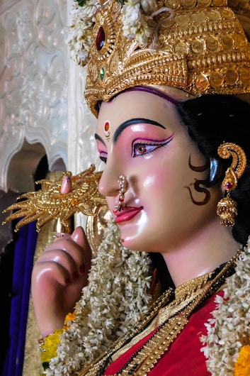A detailed sculpture of a deity adorned with an ornate golden crown embellished with jewels. The deity's face is gracefully painted with vibrant colors, featuring a bindi on the forehead. Floral garlands drape around the neck, and the figure holds a decorative object in hand. Intricate jewelry highlights the divinity's serene expression.