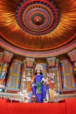 A vibrant and intricately designed temple interior featuring a richly adorned statue of a deity seated on a throne. The figure wears a blue garment with gold accents and holds traditional attributes in multiple hands. Surrounding the deity are colorful columns and elaborate patterns. The ceiling above is ornately decorated with concentric circles of diverse colors, enhancing the spiritual ambiance.