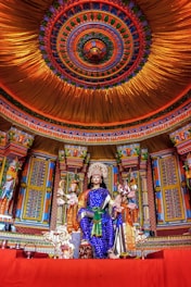 A vibrant and intricately designed temple interior featuring a richly adorned statue of a deity seated on a throne. The figure wears a blue garment with gold accents and holds traditional attributes in multiple hands. Surrounding the deity are colorful columns and elaborate patterns. The ceiling above is ornately decorated with concentric circles of diverse colors, enhancing the spiritual ambiance.
