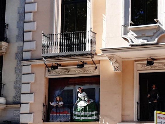 A building facade with decorative elements including a mural or painted figures resembling historical or classical attire and styles. The characters appear to be standing on a small balcony. Architectural details such as wrought iron railing, and ornate window frames are visible.