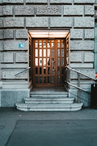 An entrance with a wooden door featuring multiple glass panels is flanked by two metal railings. It is set within a stone wall with large, textured blocks. The entrance is elevated by a few steps leading to the doorway, and a small blue sign with the number three is visible on the left.