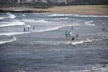 Several people are engaged in surfing activities on a beach with gentle waves. The ocean is slightly choppy with visible whitecaps. Surfers are either on their boards riding waves or carrying their boards in the surf. The background shows a sandy beach and a residential area with multiple buildings along the shoreline.
