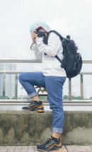 A person wearing a light blue helmet, white sweater, and rolled-up blue jeans holds a camera, seemingly taking a photo. They have a large black backpack and are standing next to a railing with a cityscape and body of water in the background.