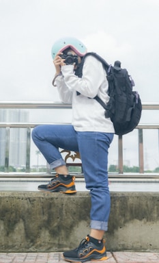 A person wearing a light blue helmet, white sweater, and rolled-up blue jeans holds a camera, seemingly taking a photo. They have a large black backpack and are standing next to a railing with a cityscape and body of water in the background.