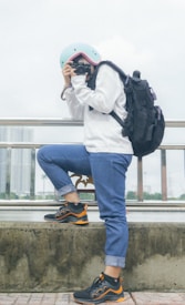 A person wearing a light blue helmet, white sweater, and rolled-up blue jeans holds a camera, seemingly taking a photo. They have a large black backpack and are standing next to a railing with a cityscape and body of water in the background.