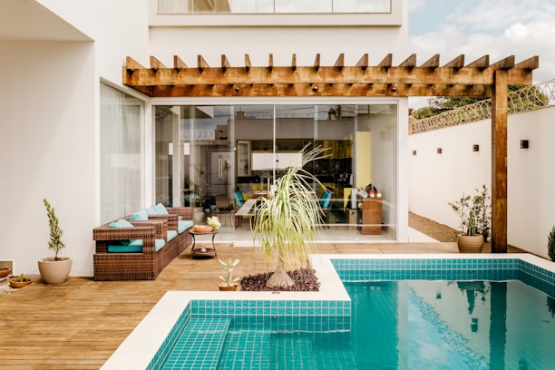 A modern outdoor patio area featuring a wooden pergola over a spacious wicker lounge set with teal cushions. A small swimming pool with clear blue tiles is surrounded by a wooden deck. Various potted plants, including a large decorative grass and small shrubs, add greenery. The space is enclosed by tall walls and a large glass sliding door leads into an interior room that can be seen in the background.