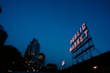 A neon sign reading 'Public Market' stands prominently against a deep blue evening sky. The sign is mounted on a metal framework and is illuminated, casting a warm glow. In the background, a tall city building displays its lights, creating an urban skyline.