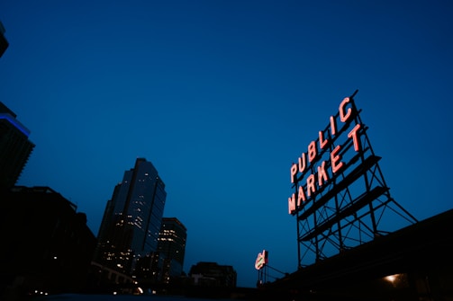 A neon sign reading 'Public Market' stands prominently against a deep blue evening sky. The sign is mounted on a metal framework and is illuminated, casting a warm glow. In the background, a tall city building displays its lights, creating an urban skyline.