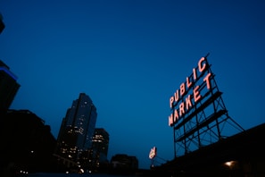 A neon sign reading 'Public Market' stands prominently against a deep blue evening sky. The sign is mounted on a metal framework and is illuminated, casting a warm glow. In the background, a tall city building displays its lights, creating an urban skyline.