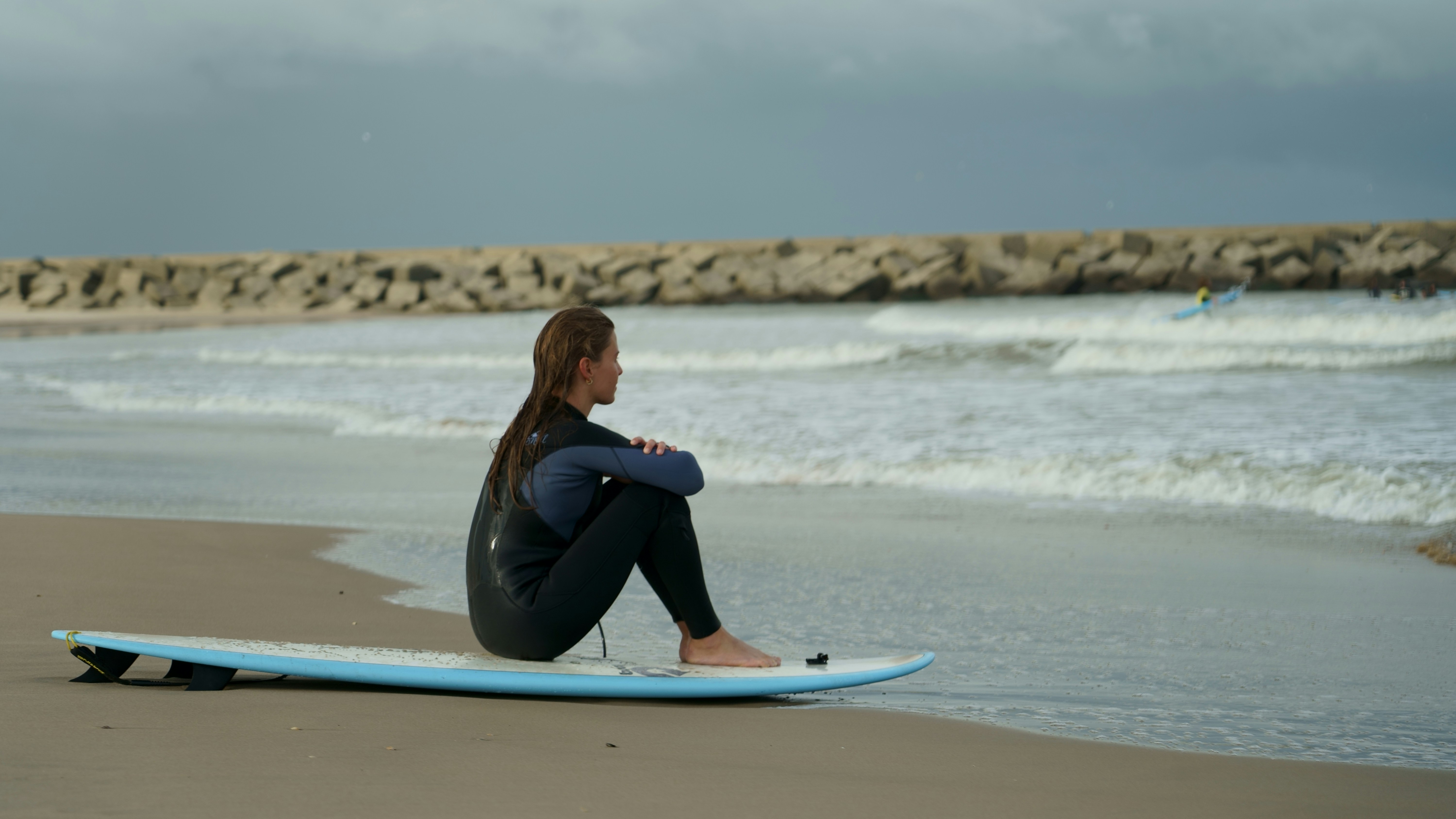 donna in camicia blu a maniche lunghe e pantaloni neri seduta sulla tavola da surf blu sulla spiaggia durante