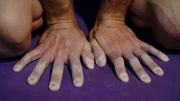 Two hands placed on a purple mat, with fingers spread wide. The hands appear to be supporting bodyweight, slightly tensed, suggesting a yoga pose or exercise. The skin on the hands is slightly chalky, possibly due to the use of gym chalk for grip.