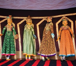 Four intricately designed Rajasthani puppets hang against a colorful backdrop. Each puppet is adorned in traditional attire with vibrant patterns and detailed facial features. They are dressed in long gowns with elaborate embroidery, showcasing a rich cultural heritage.