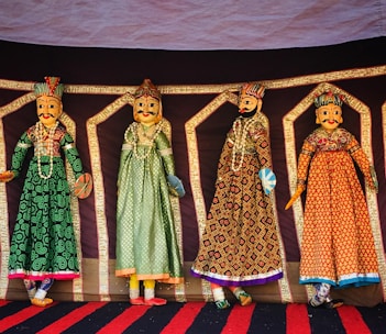 Four intricately designed Rajasthani puppets hang against a colorful backdrop. Each puppet is adorned in traditional attire with vibrant patterns and detailed facial features. They are dressed in long gowns with elaborate embroidery, showcasing a rich cultural heritage.
