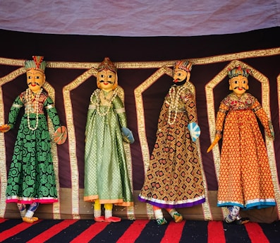 Four intricately designed Rajasthani puppets hang against a colorful backdrop. Each puppet is adorned in traditional attire with vibrant patterns and detailed facial features. They are dressed in long gowns with elaborate embroidery, showcasing a rich cultural heritage.