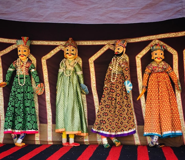 Four intricately designed Rajasthani puppets hang against a colorful backdrop. Each puppet is adorned in traditional attire with vibrant patterns and detailed facial features. They are dressed in long gowns with elaborate embroidery, showcasing a rich cultural heritage.