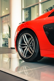 A vibrant red sports car is partially visible, focusing on the front wheel and sleek body design. The shiny alloy wheel is prominently featured, reflecting the showroom lights. The modern automotive design is emphasized by the curved lines and glossy finish.