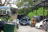 An outdoor area filled with cars, some stacked on top of each other, indicating a car junkyard or repair facility. Vehicles appear in various states of disassembly, with an open hood on the black car in the middle. A forklift is visible near the right, surrounded by green foliage and trees.