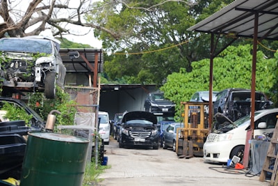 An outdoor area filled with cars, some stacked on top of each other, indicating a car junkyard or repair facility. Vehicles appear in various states of disassembly, with an open hood on the black car in the middle. A forklift is visible near the right, surrounded by green foliage and trees.
