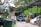 An outdoor area filled with cars, some stacked on top of each other, indicating a car junkyard or repair facility. Vehicles appear in various states of disassembly, with an open hood on the black car in the middle. A forklift is visible near the right, surrounded by green foliage and trees.